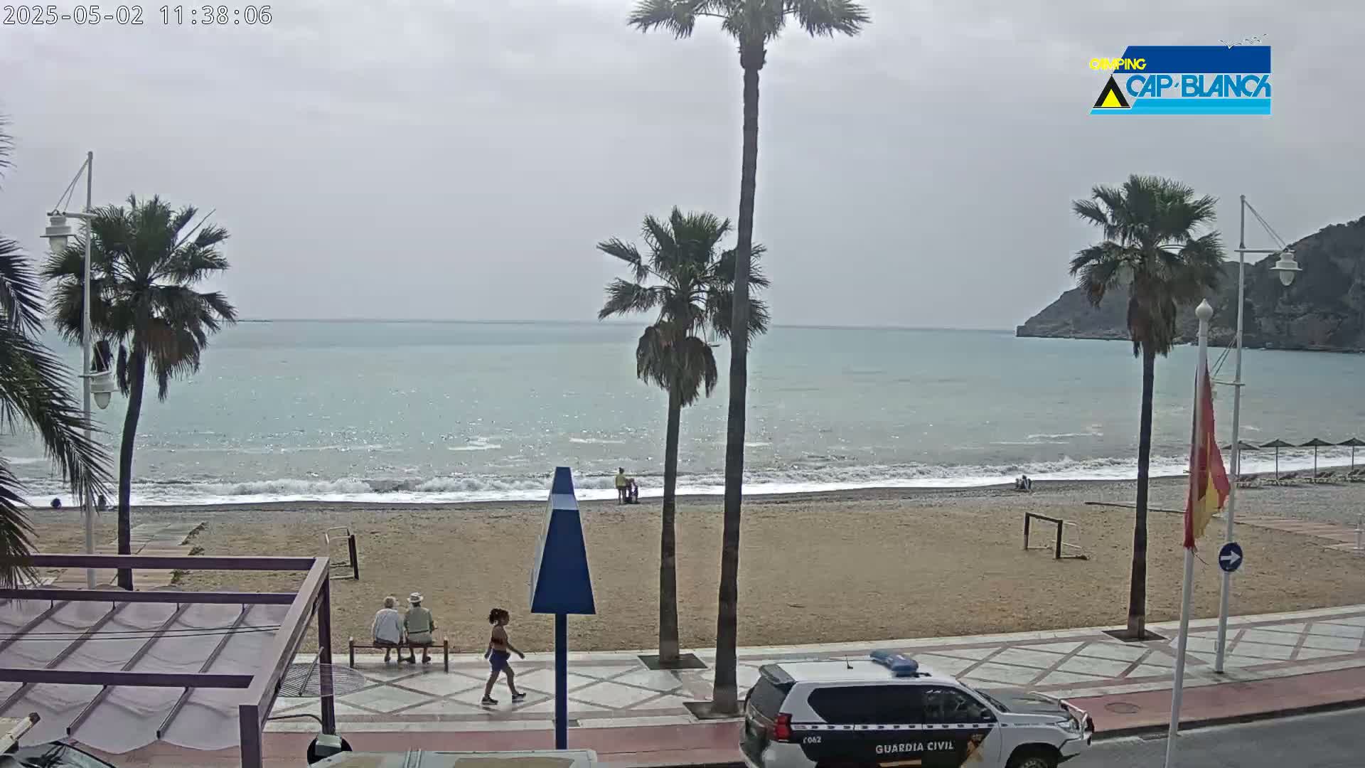 A sandy beach with calm, light-blue water under an overcast sky is seen from above, with palm trees, people walking, and a police car parked nearby.
