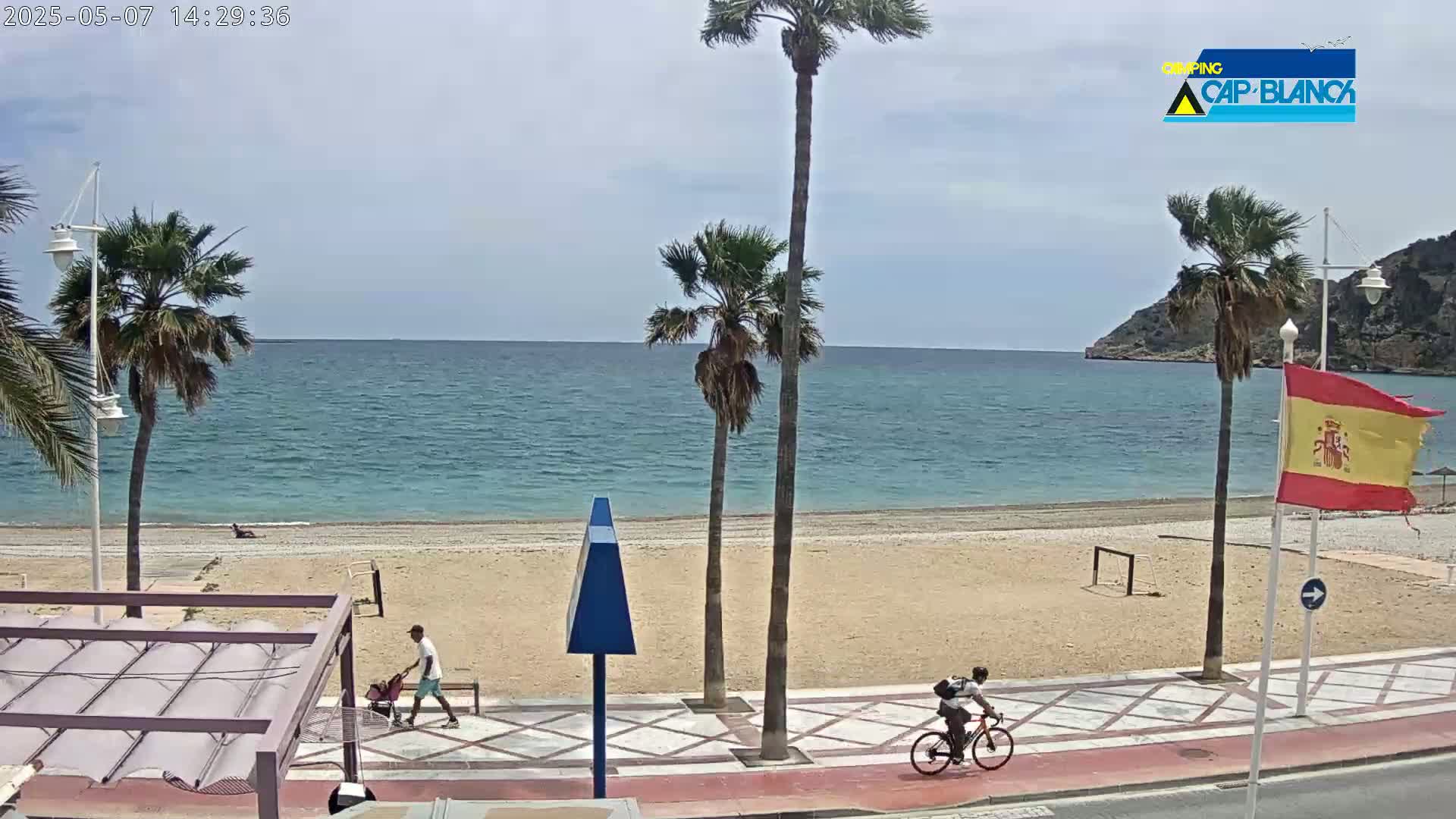 A sandy beach with calm blue water, palm trees, and a few people walking and biking, under a mostly clear sky.