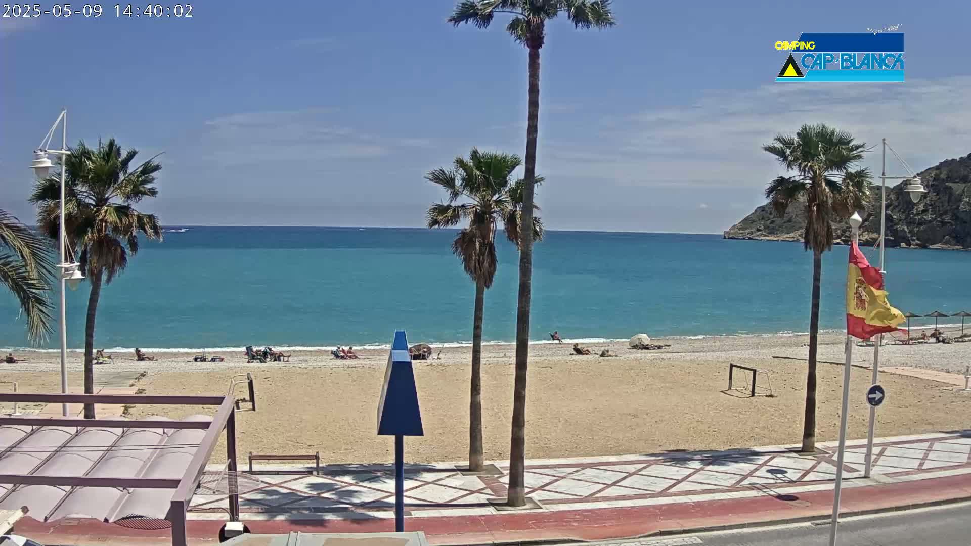 A sandy beach with a few people on it stretches along a calm, turquoise sea under a clear blue sky, lined with palm trees and a paved walkway.