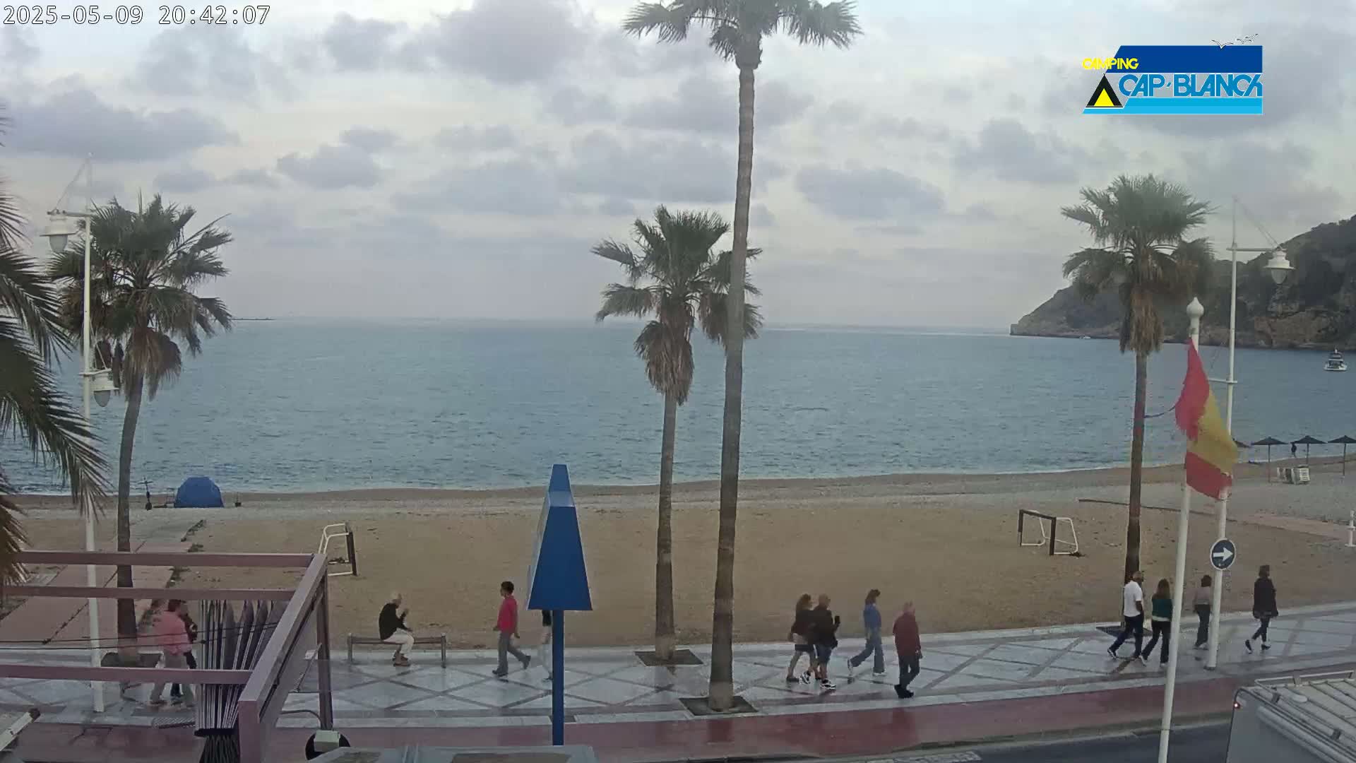 A mostly cloudy day reveals a sandy beach with palm trees and several people walking along a promenade next to a calm sea.