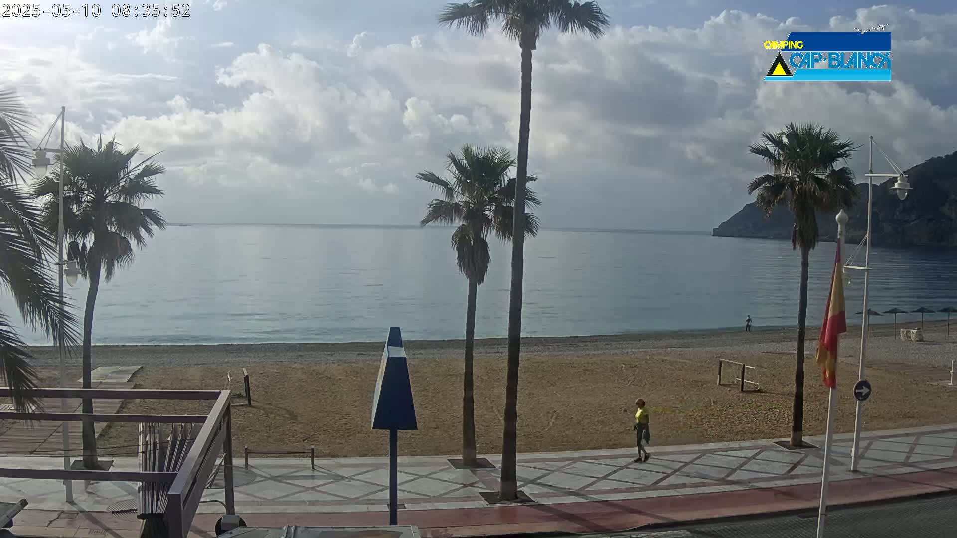 A partly cloudy morning scene shows a sandy beach next to a calm ocean, bordered by a paved walkway and several palm trees.