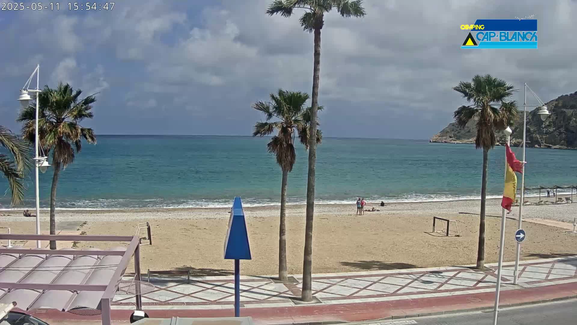 A sandy beach with palm trees and a few people on it, next to a calm turquoise ocean under a partly cloudy sky.
