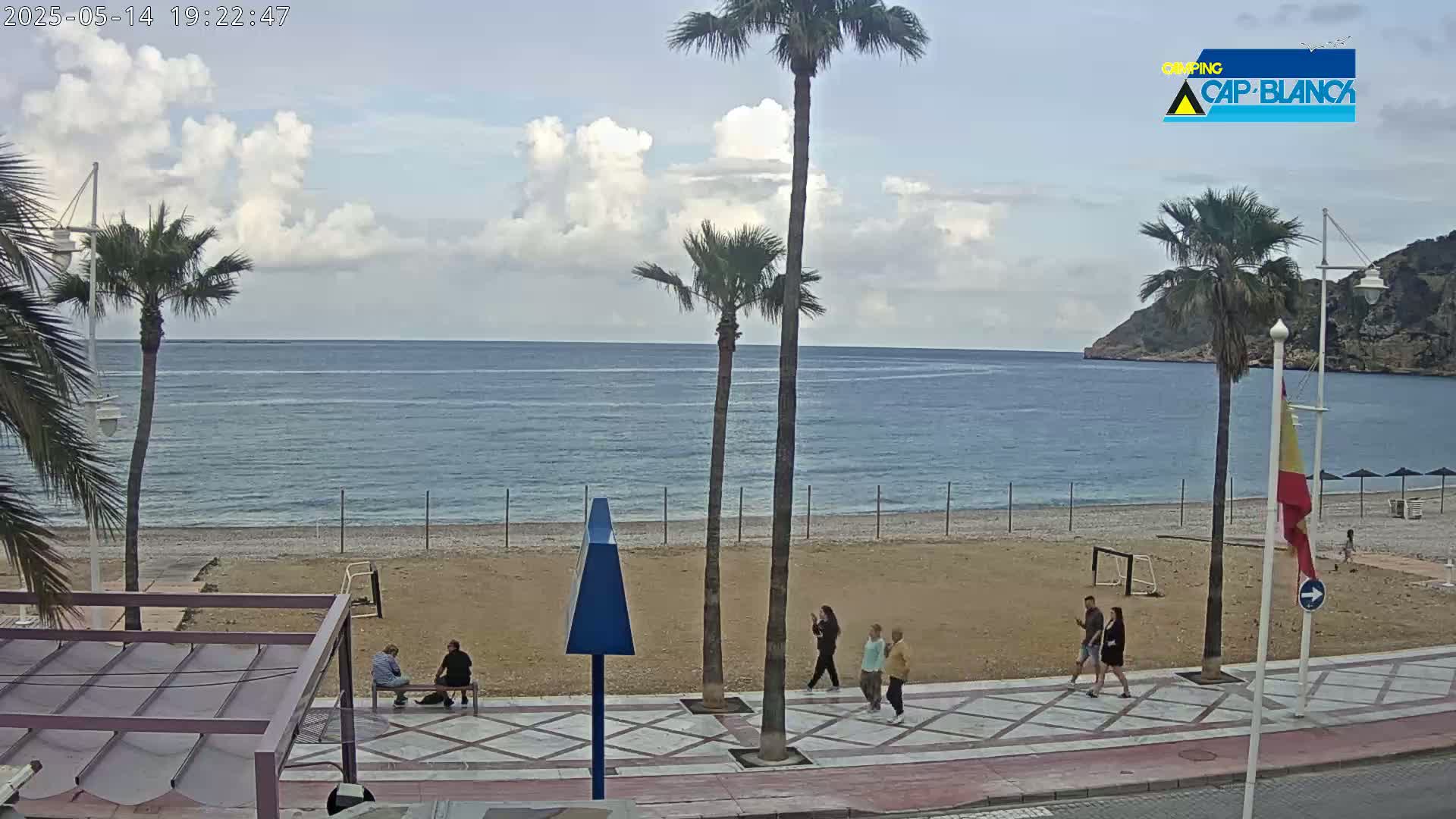 A mostly sunny beach scene with a calm sea, palm trees, and a few people walking along a paved walkway.