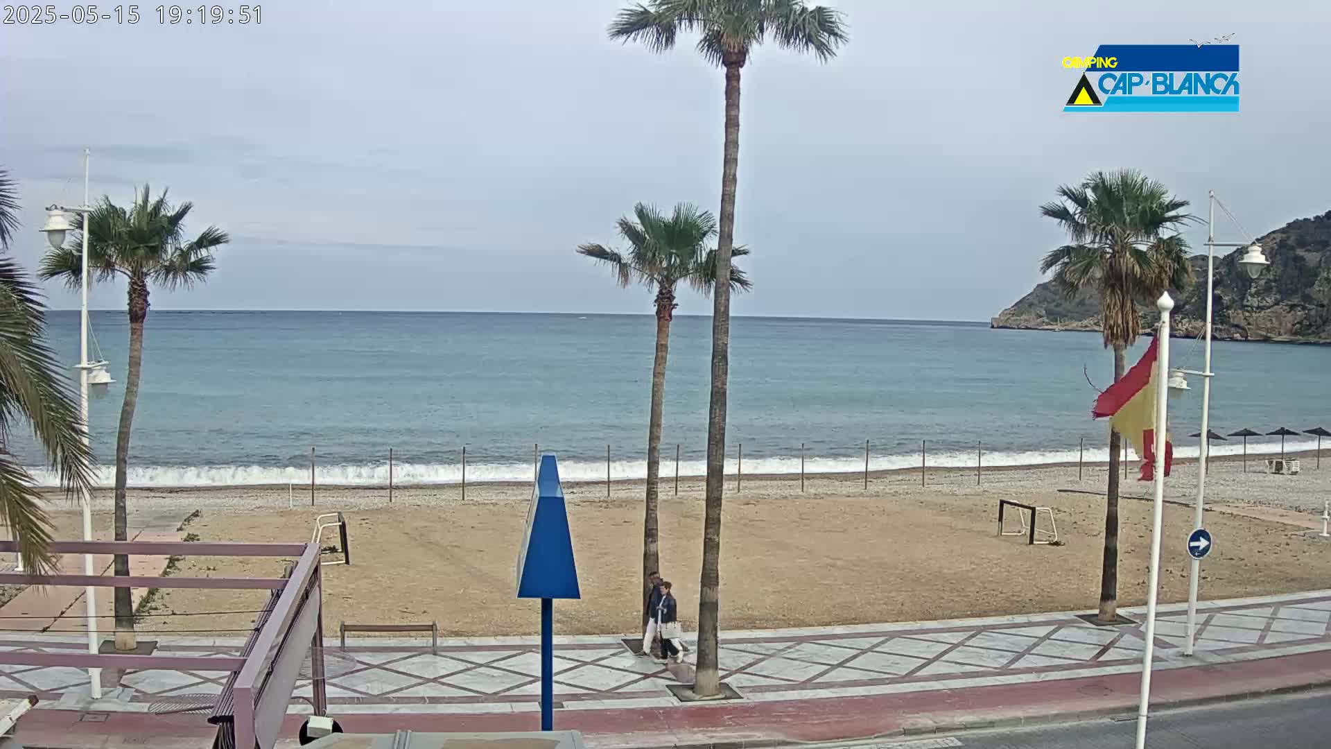 A mostly clear, overcast day at a beach with calm waves, palm trees, and two people walking along the promenade.
