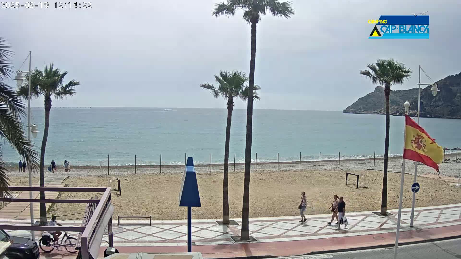 A mostly sunny beach scene shows a sandy beach with a calm ocean, palm trees, people walking on a promenade, and a Spanish flag.