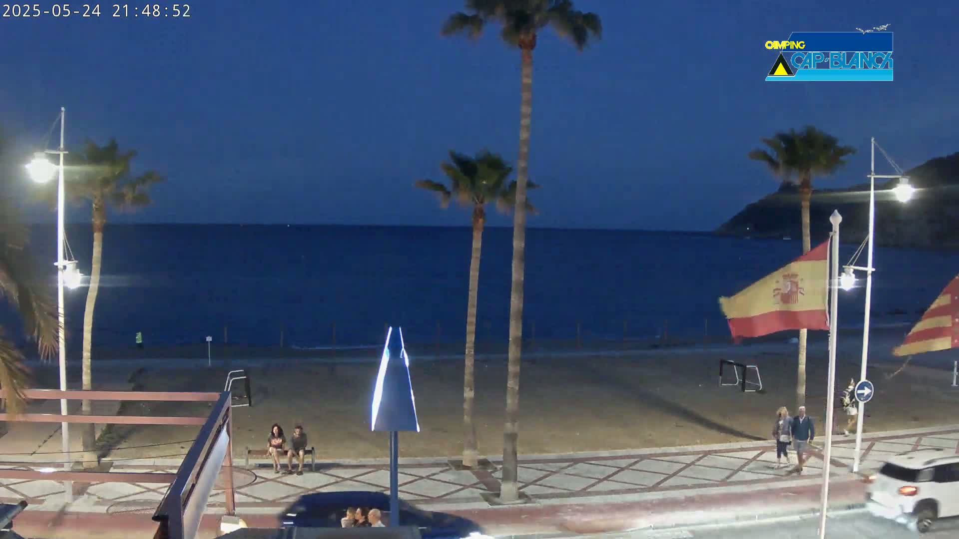 A nighttime beach scene shows a calm, dark-blue sea under a twilight sky, with palm trees, people walking along a paved walkway, and a few cars parked nearby.
