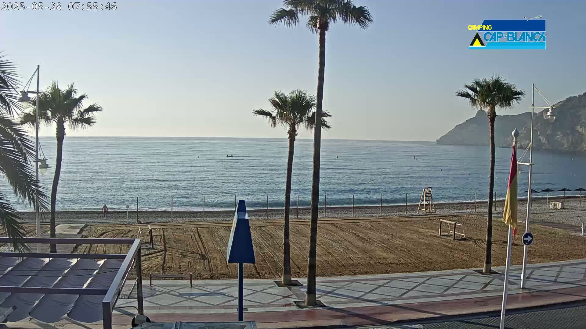 A mostly sunny beach with calm ocean waters, palm trees, and an empty volleyball court is visible from a boardwalk.