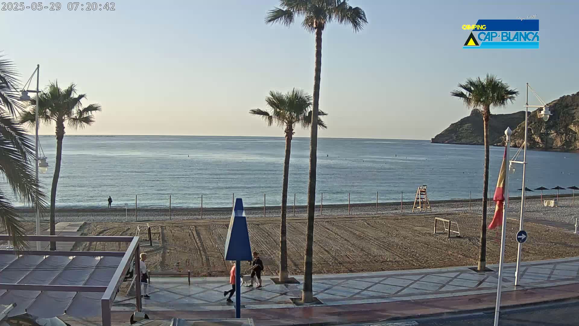A calm, bright beach scene shows a sandy beach with palm trees, a boardwalk, and a few people walking, all under a clear sky.