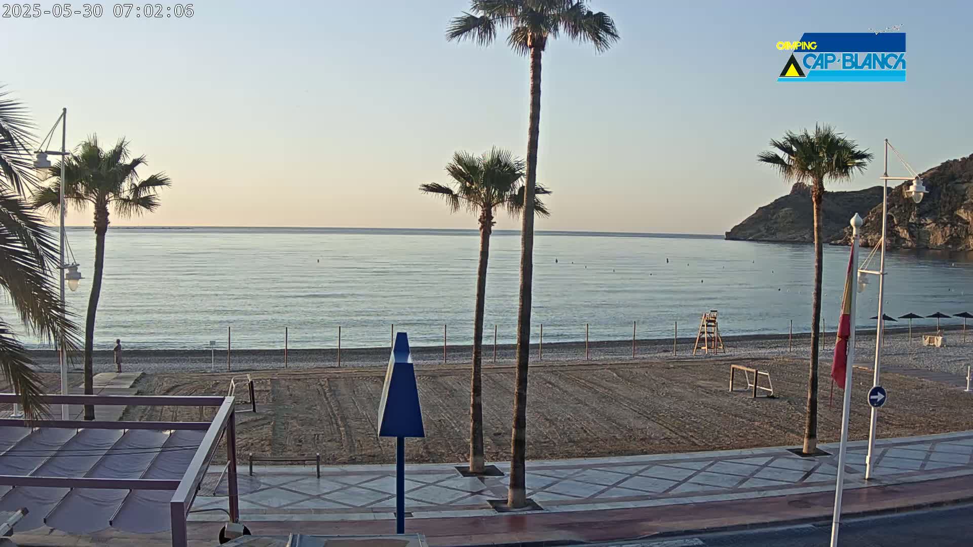 A calm, pale-blue sea stretches behind a sandy beach lined with palm trees and a boardwalk under a clear, dawn sky.