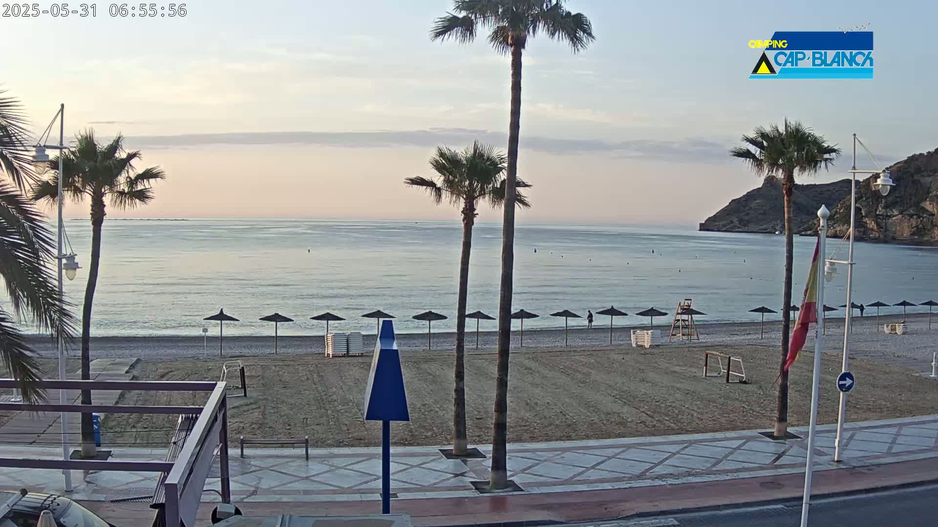 A calm, pale-blue sea stretches to a sandy beach lined with closed umbrellas at sunrise, backed by a palm tree-lined promenade and a rocky hill.