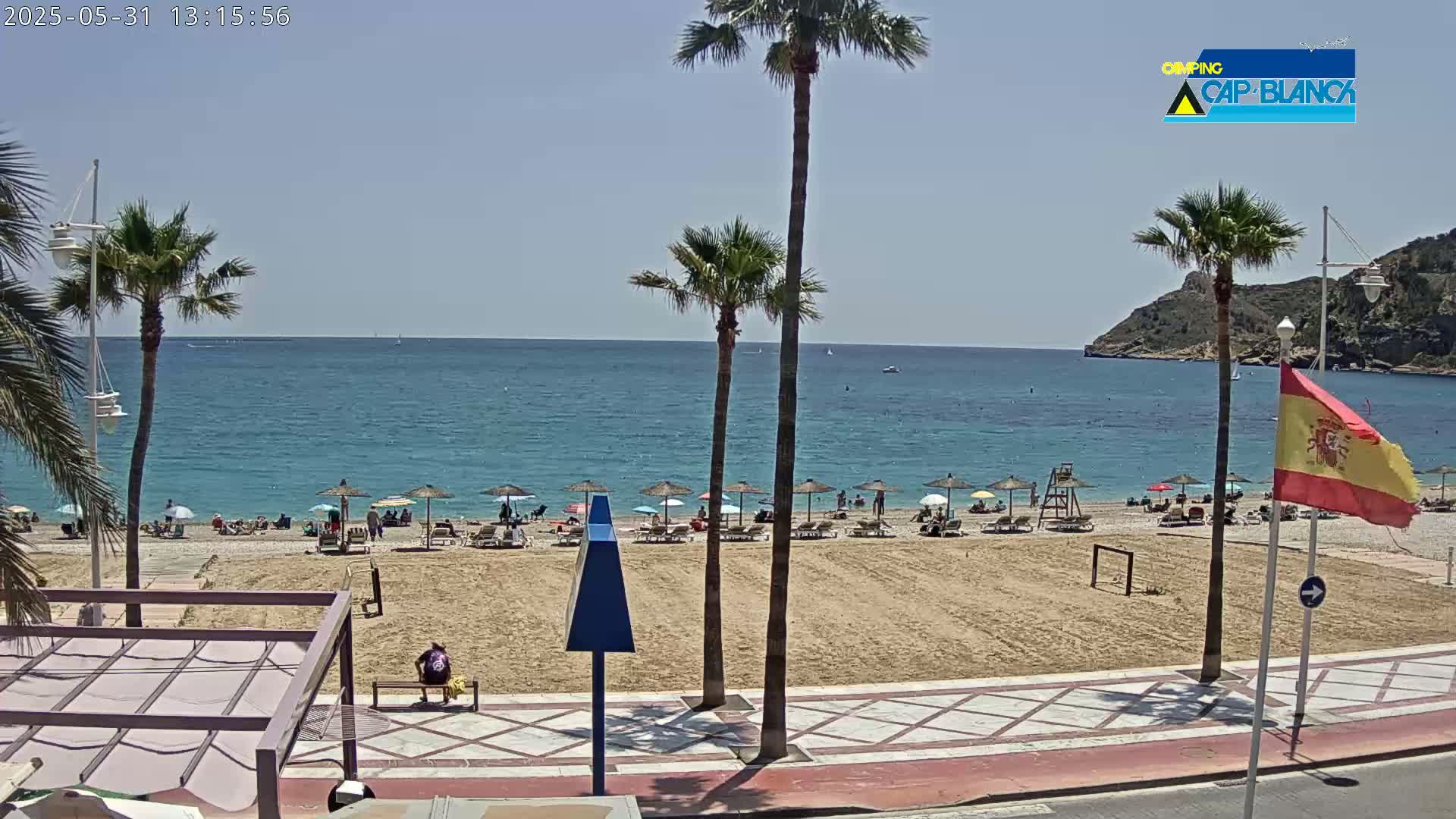 A sandy beach with numerous umbrellas and people relaxing is situated next to a calm, blue ocean under a clear sky.