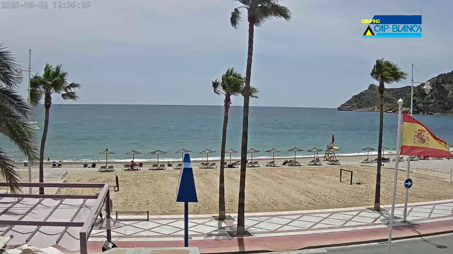 A sandy beach with palm trees and several beach umbrellas is visible, alongside a calm ocean under a mostly clear sky.