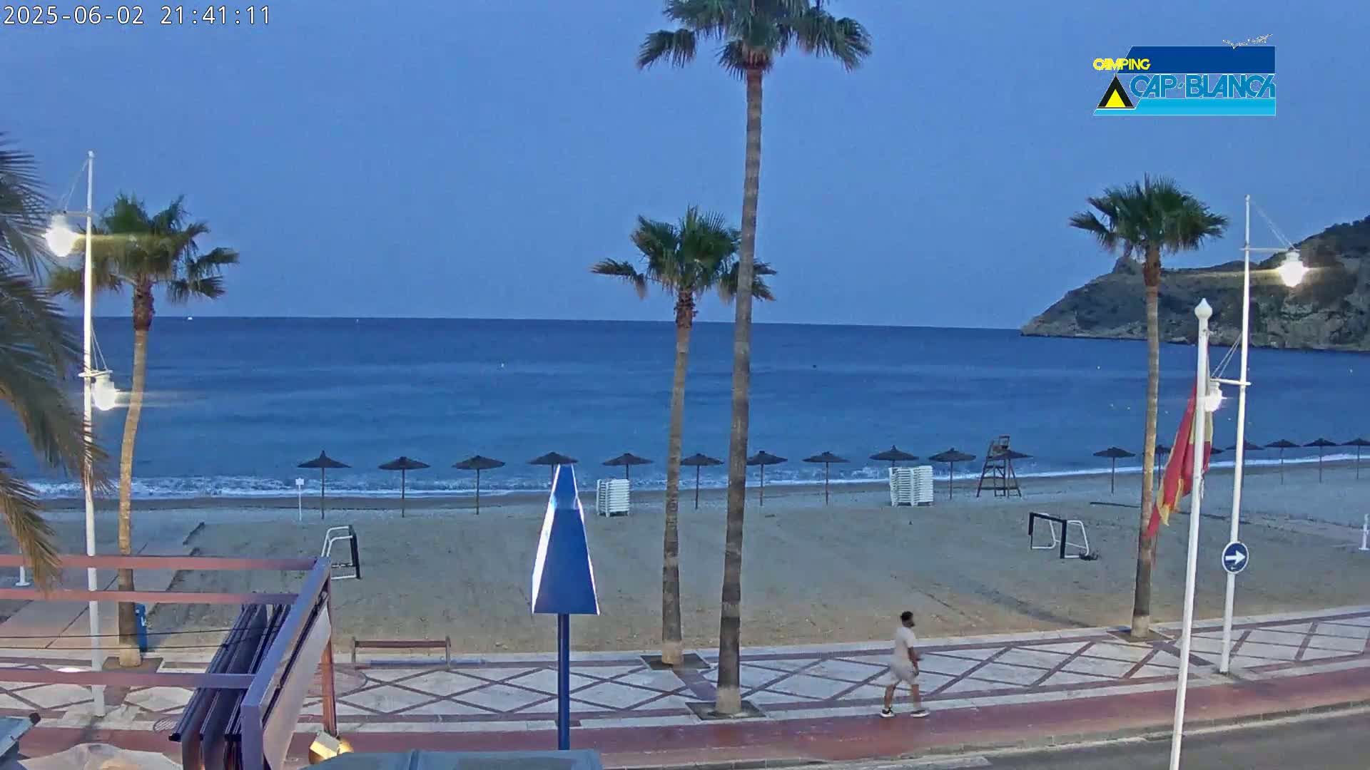 A mostly empty sandy beach at dusk, with palm trees lining a paved walkway and a calm blue sea in the background.