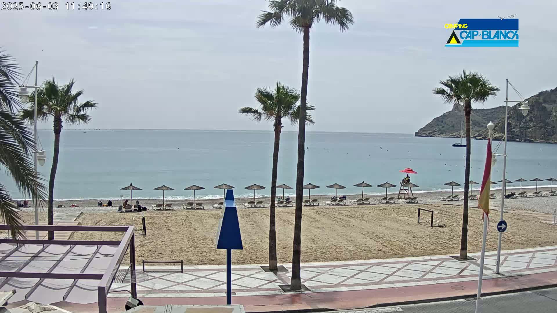 A sandy beach with several beach umbrellas and lounge chairs is visible, overlooking a calm ocean under a mostly cloudy sky.