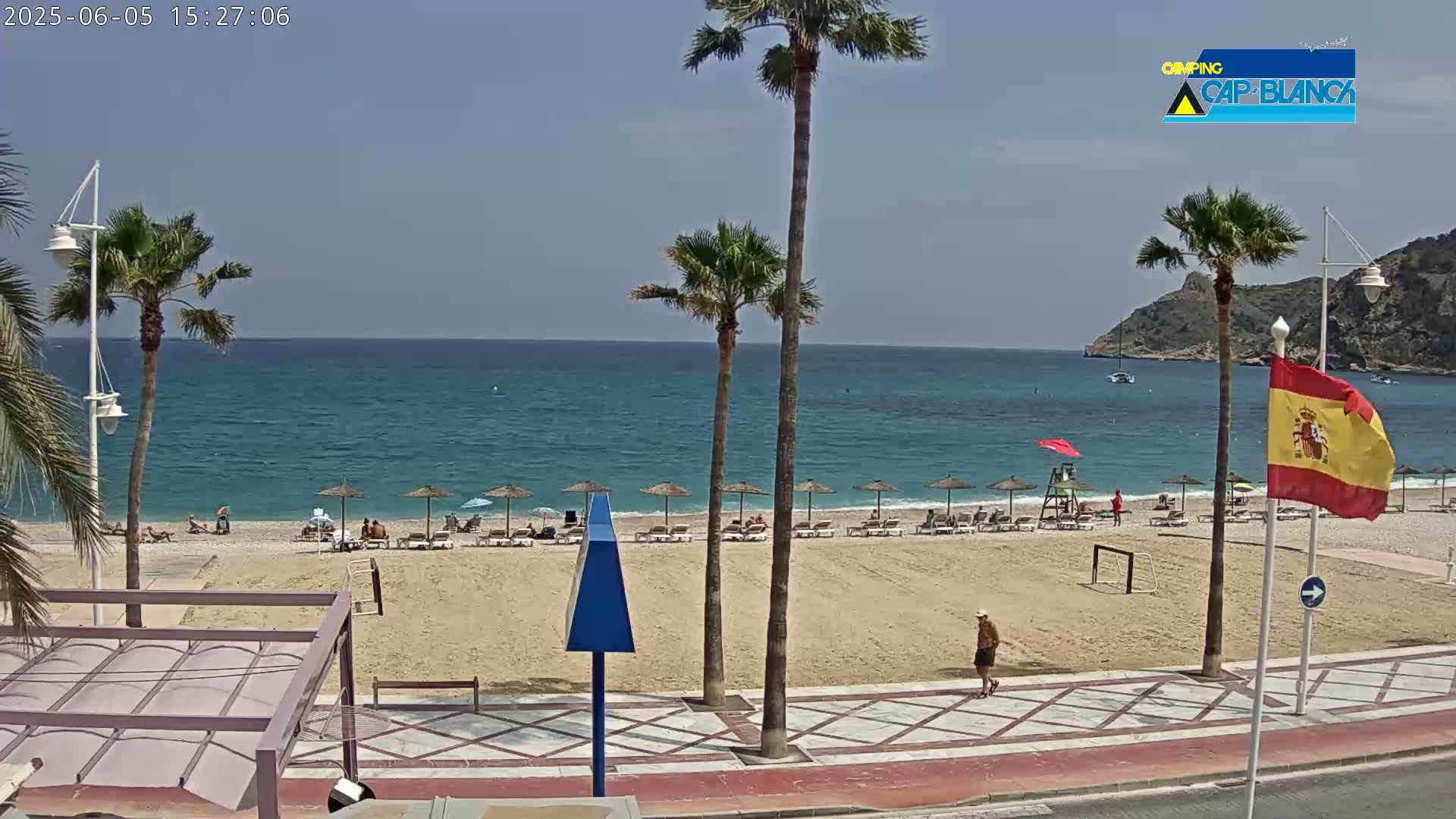A sunny beach scene shows a sandy shore with scattered beach umbrellas and lounge chairs, palm trees lining a paved walkway next to the beach, and a calm blue ocean stretching to a distant mountain.