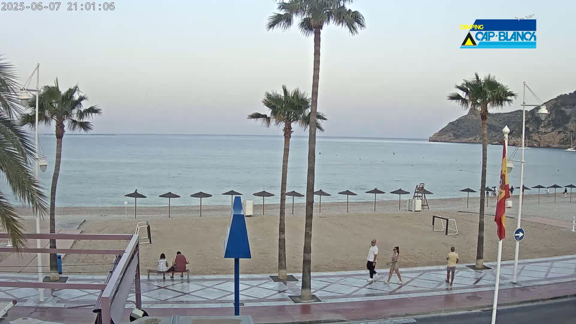A mostly empty sandy beach with calm, light blue water is bordered by a paved walkway where several people are walking, and palm trees stand near the beach.