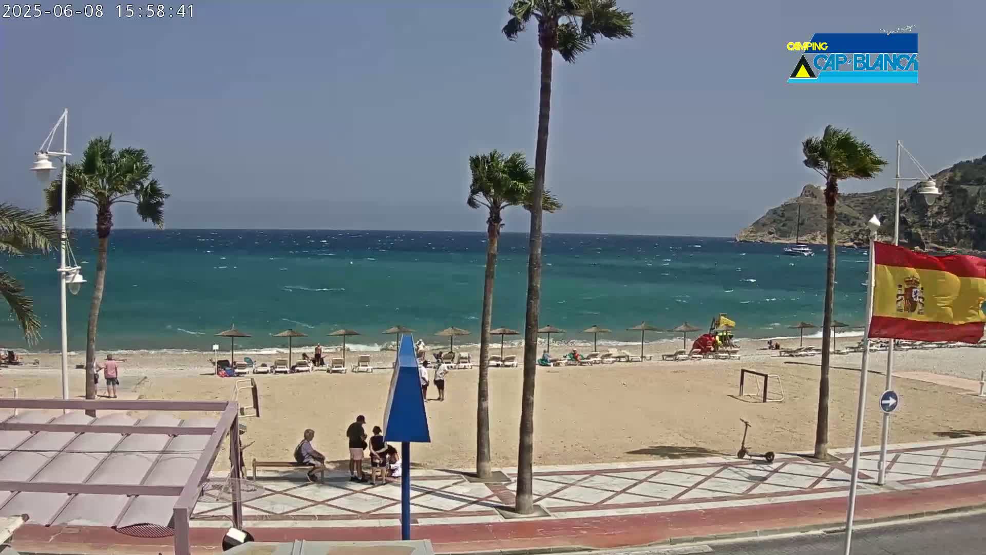 A sandy beach with scattered beach umbrellas and people, palm trees, and a partly cloudy sky over a windy turquoise sea.