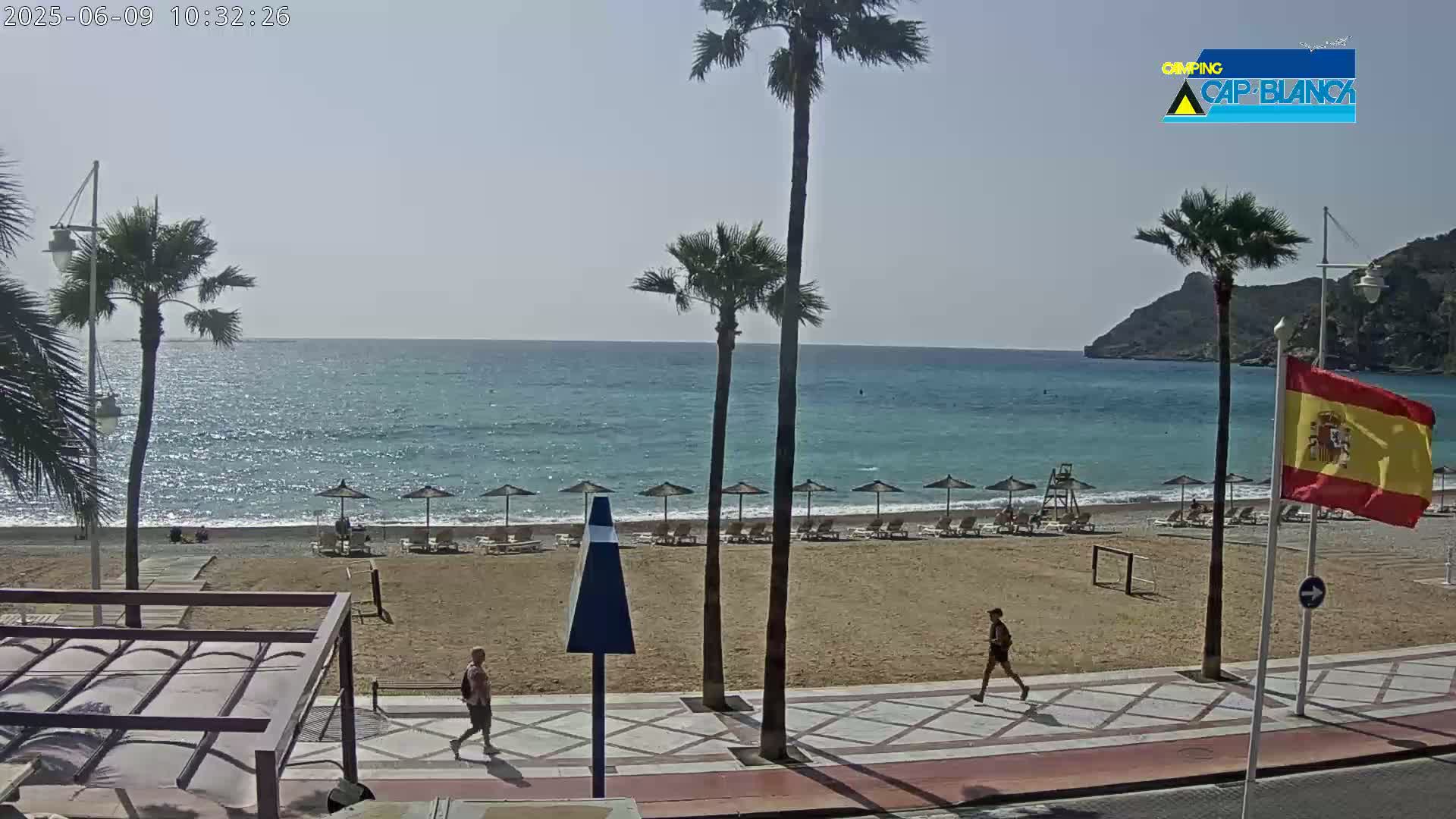 A sandy beach with calm, blue water, several palm trees, and a few people walking along a paved walkway;  the weather is sunny.