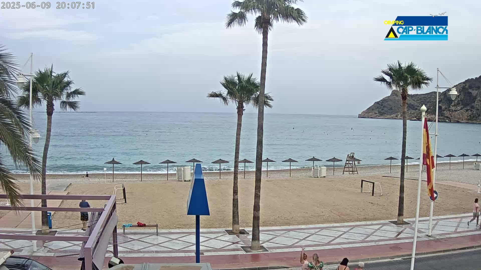 A sandy beach with palm trees and a few beach umbrellas is visible, under an overcast sky, next to a calm sea with a rocky cliff in the background.