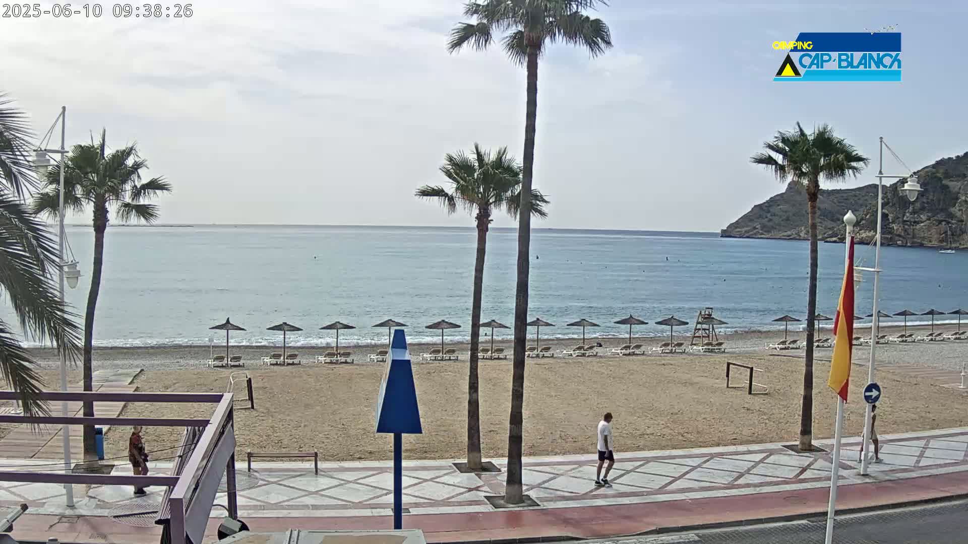 A sandy beach with calm ocean water and a few people walking along a promenade, under a partly cloudy sky.