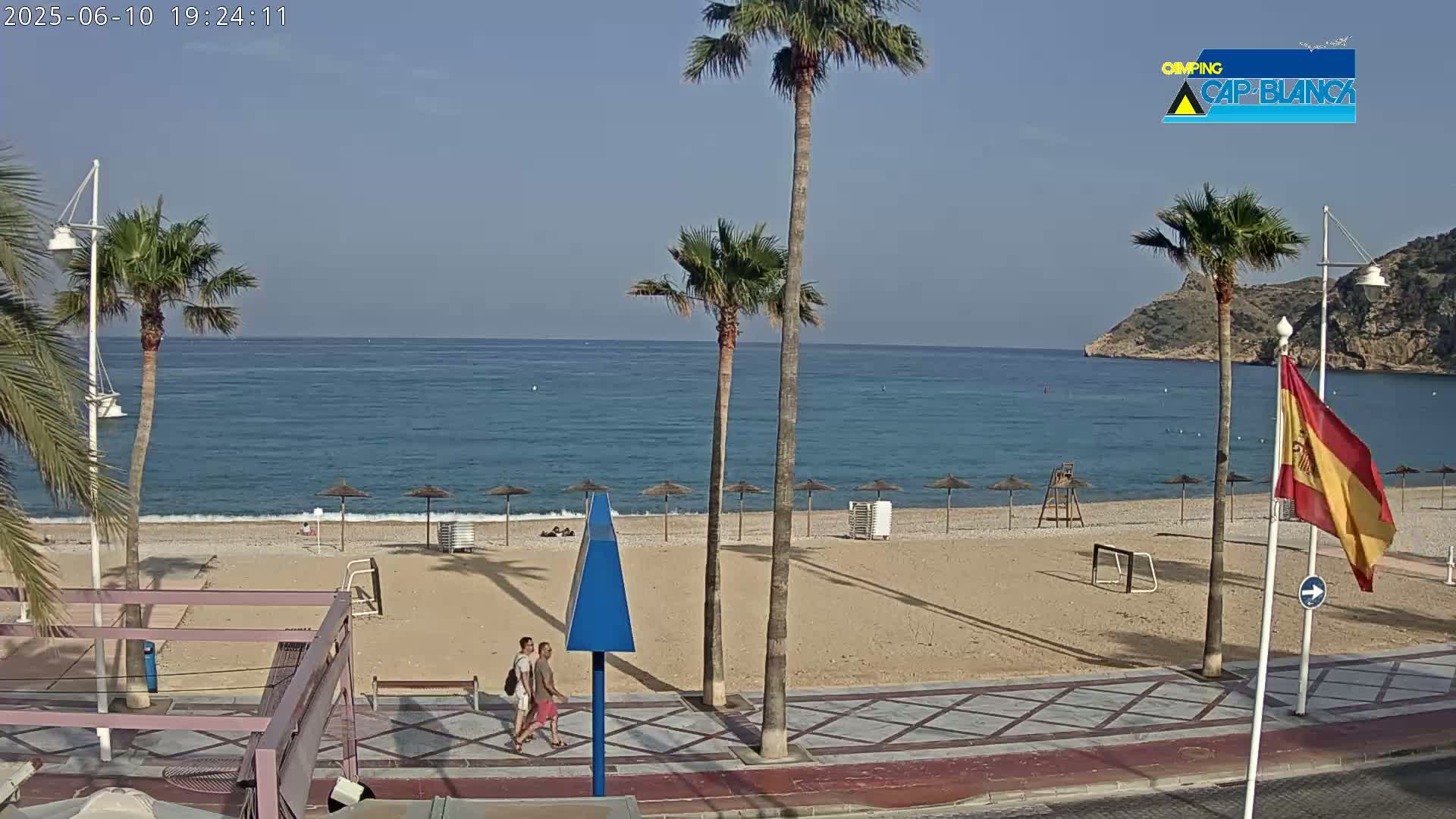 A sandy beach with palm trees, a calm ocean, and two people walking along a boardwalk on a clear, sunny day.