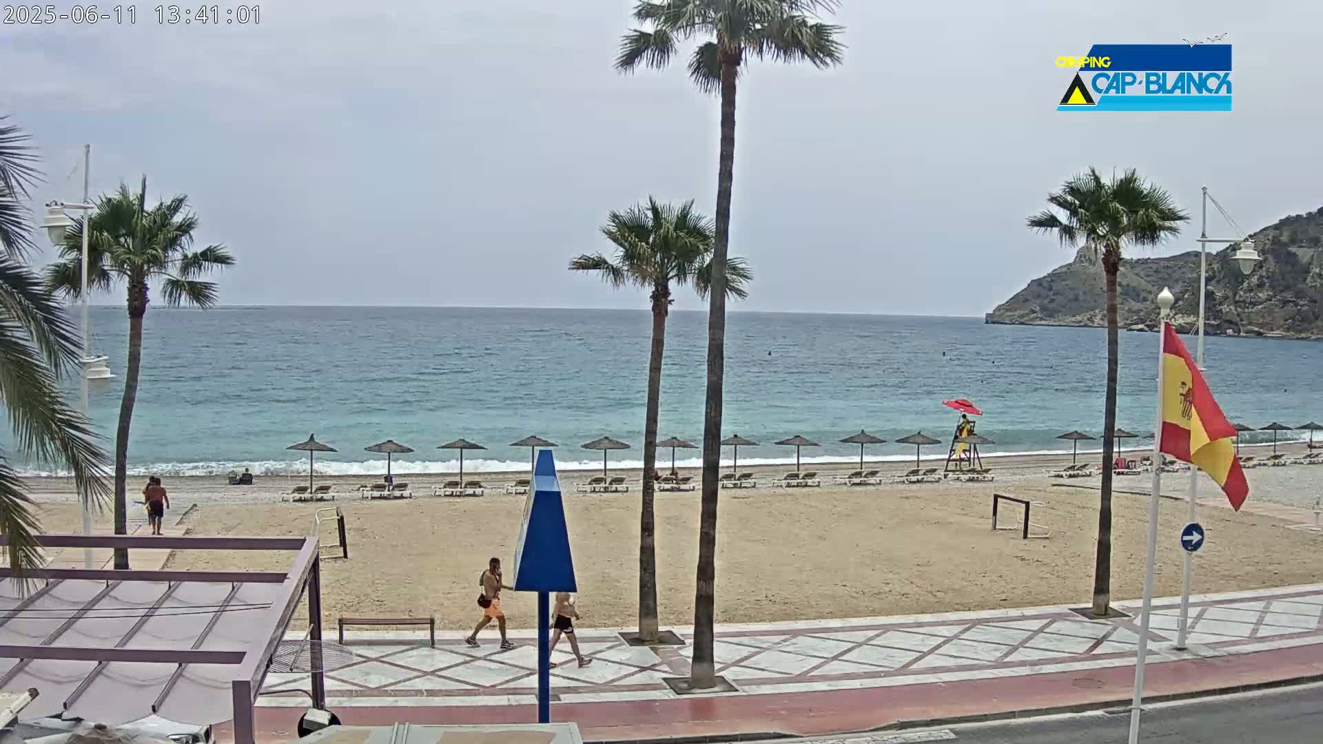 A sandy beach with several beach umbrellas and lounge chairs lines a calm, blue ocean under a partly cloudy sky, with palm trees and people walking along a paved walkway.