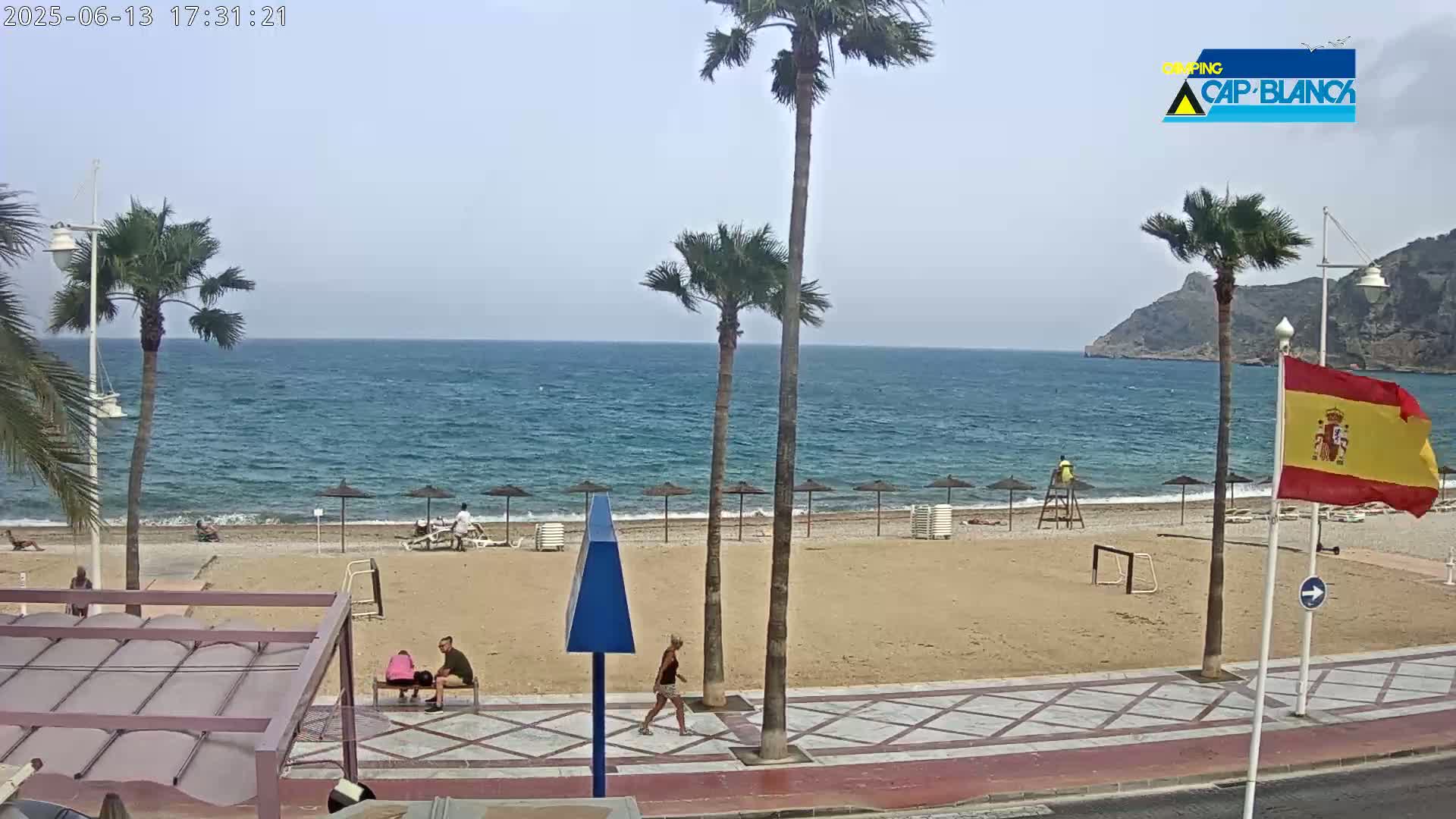 A sandy beach with palm trees, umbrellas, and a few people is visible, with a calm sea in the background under a mostly clear sky.