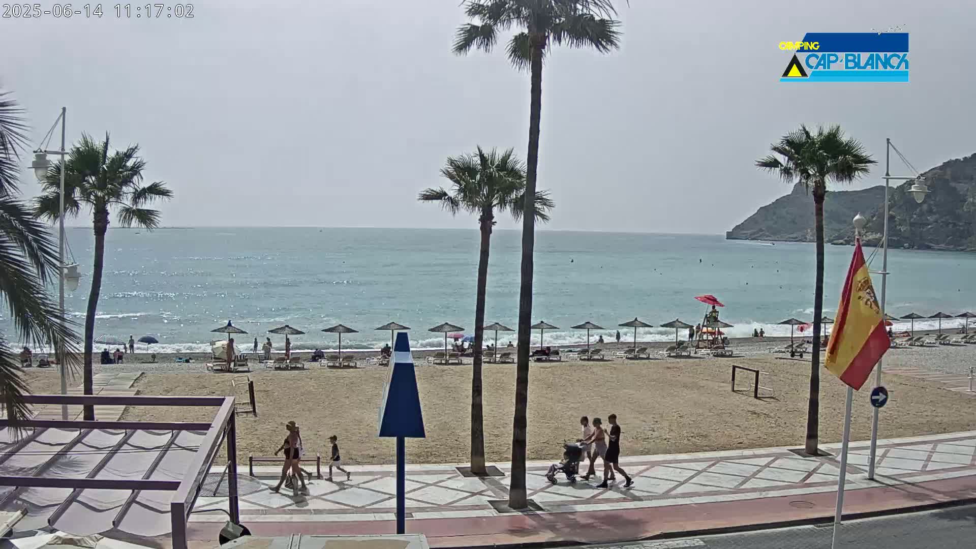 A sandy beach with calm, blue water is seen under a mostly sunny sky, with palm trees, people walking on the boardwalk, and beach umbrellas lining the shore.