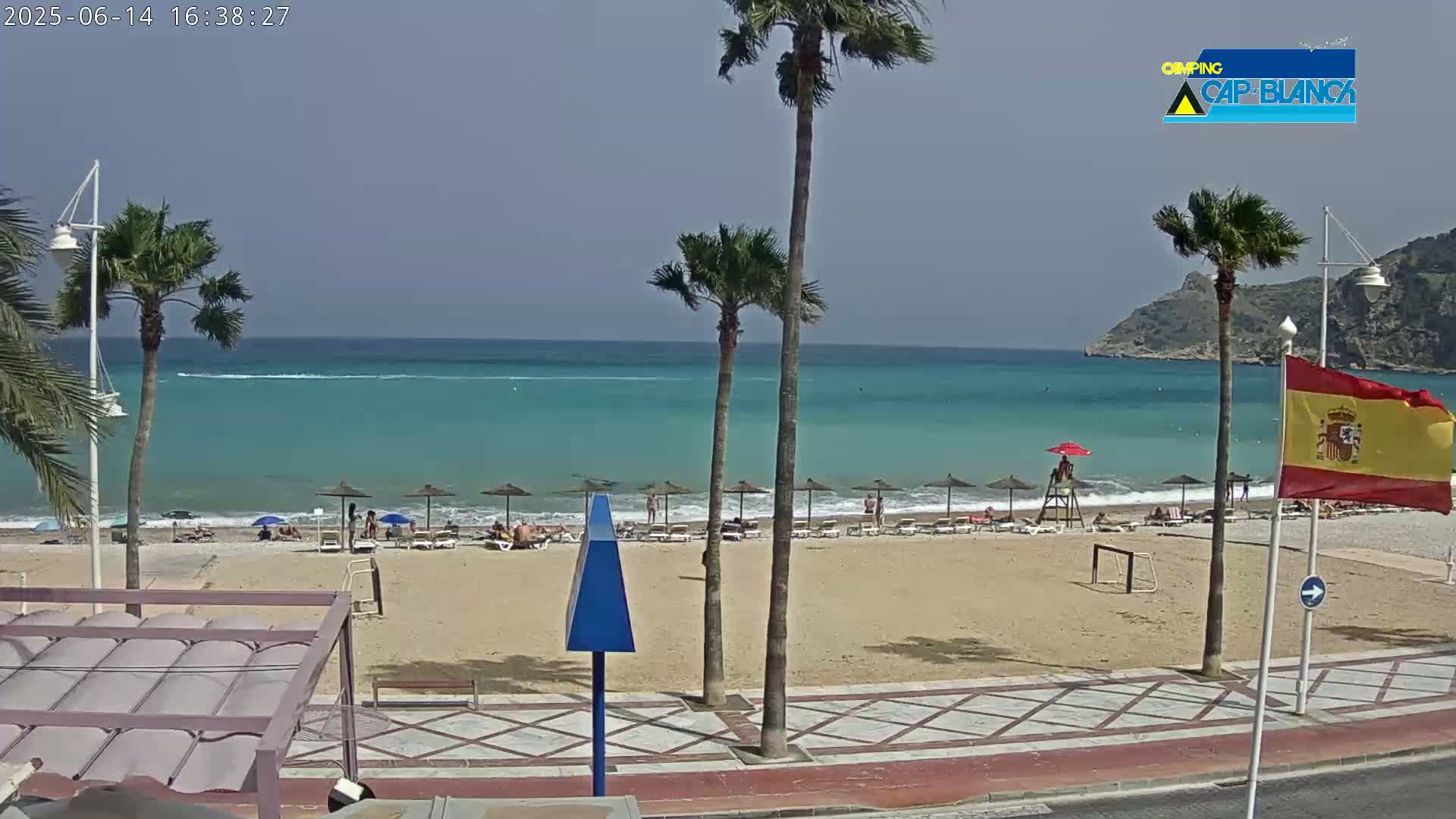 A sandy beach with turquoise water and palm trees under a mostly clear sky, showing a lifeguard stand and several beach umbrellas.