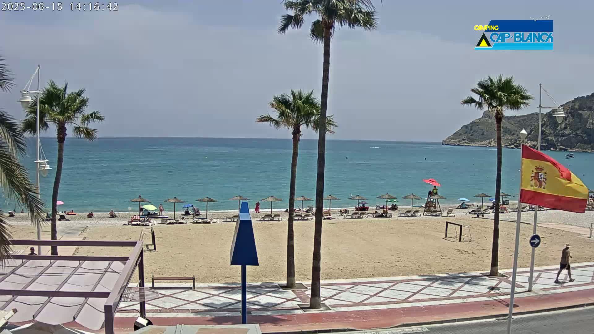 A sandy beach with several umbrellas and people, palm trees, and a calm blue ocean under a mostly clear sky is visible from a seaside promenade.