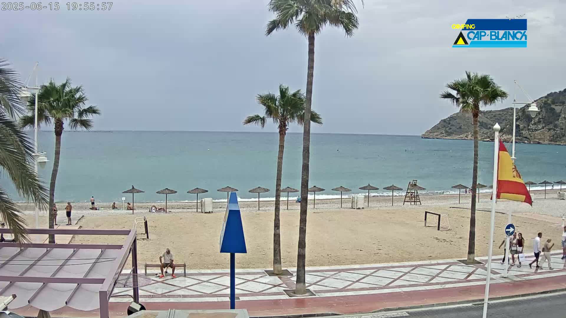 A sandy beach with several people and beach umbrellas is visible, under an overcast sky, next to calm, light-blue ocean water and a rocky cliff in the background.