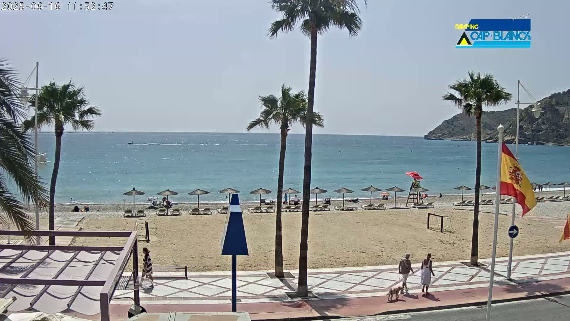 A sandy beach with several umbrellas and lounge chairs, palm trees, and a few people walking along a promenade next to a calm, blue ocean on a sunny day.