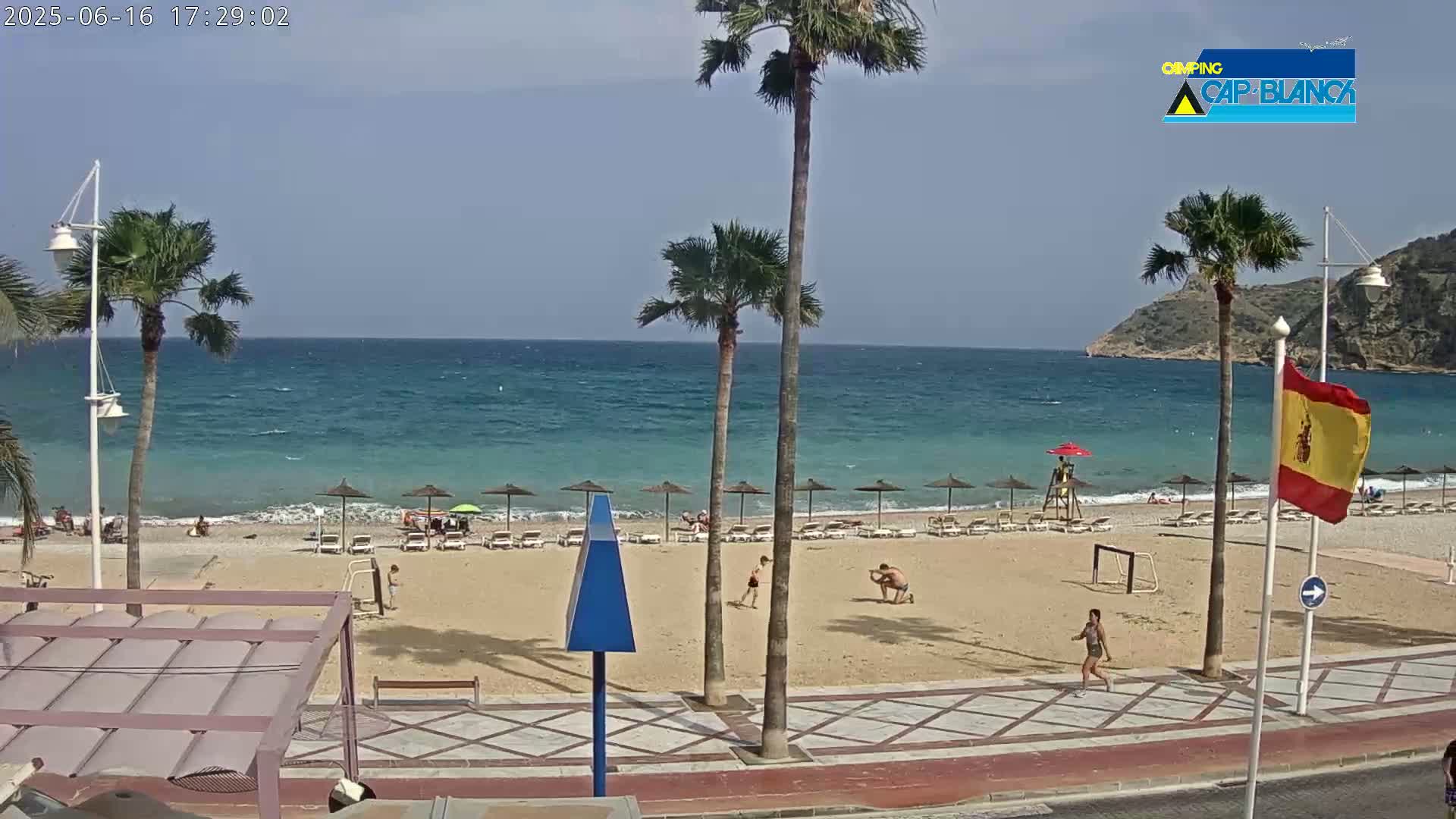 A sandy beach with palm trees, beach umbrellas, and a few people is visible, overlooking a calm, blue ocean under a clear sky.