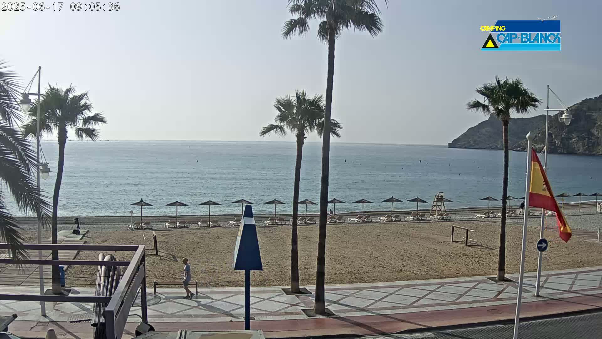 A sandy beach with a few people and beach umbrellas stretches along a calm ocean, backed by palm trees and a paved walkway under a clear, bright sky.