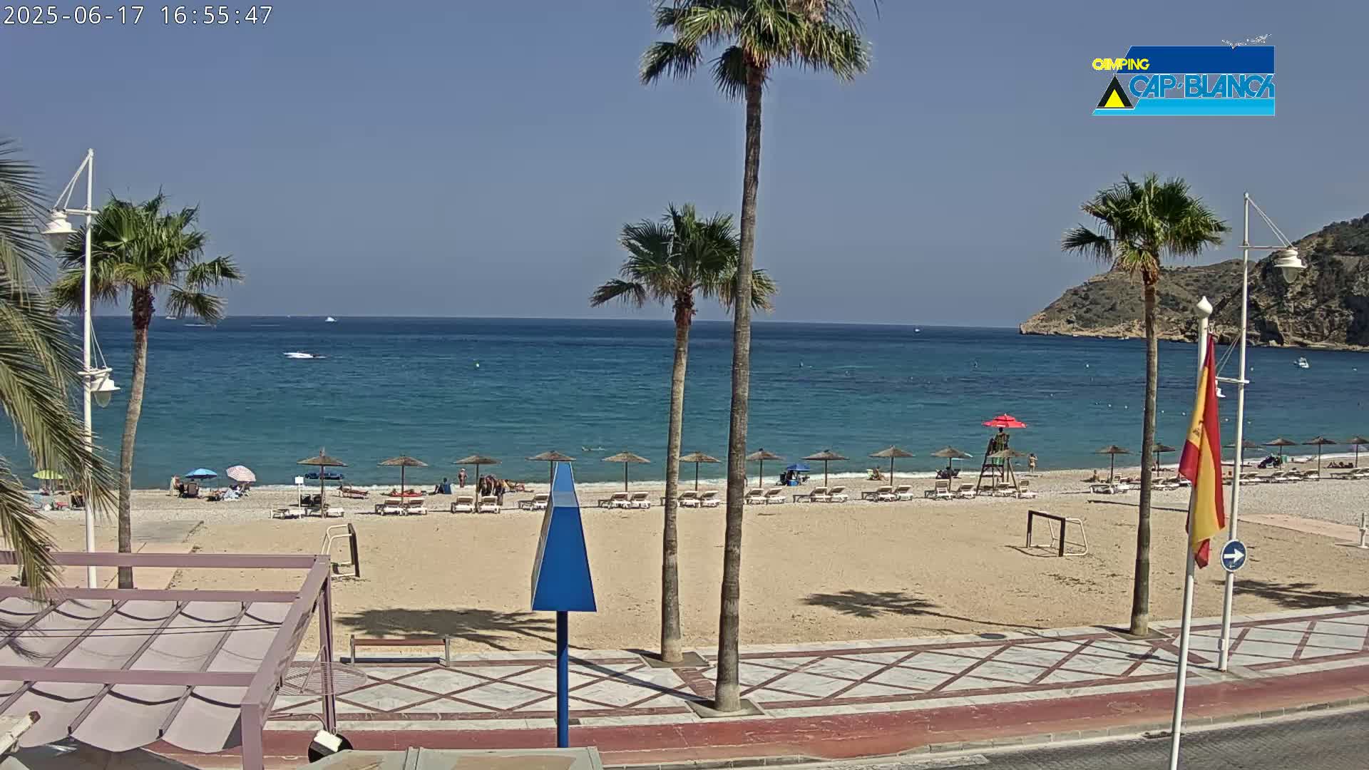 A sandy beach with several people and umbrellas, bordered by palm trees and a calm, blue ocean under a clear sky.