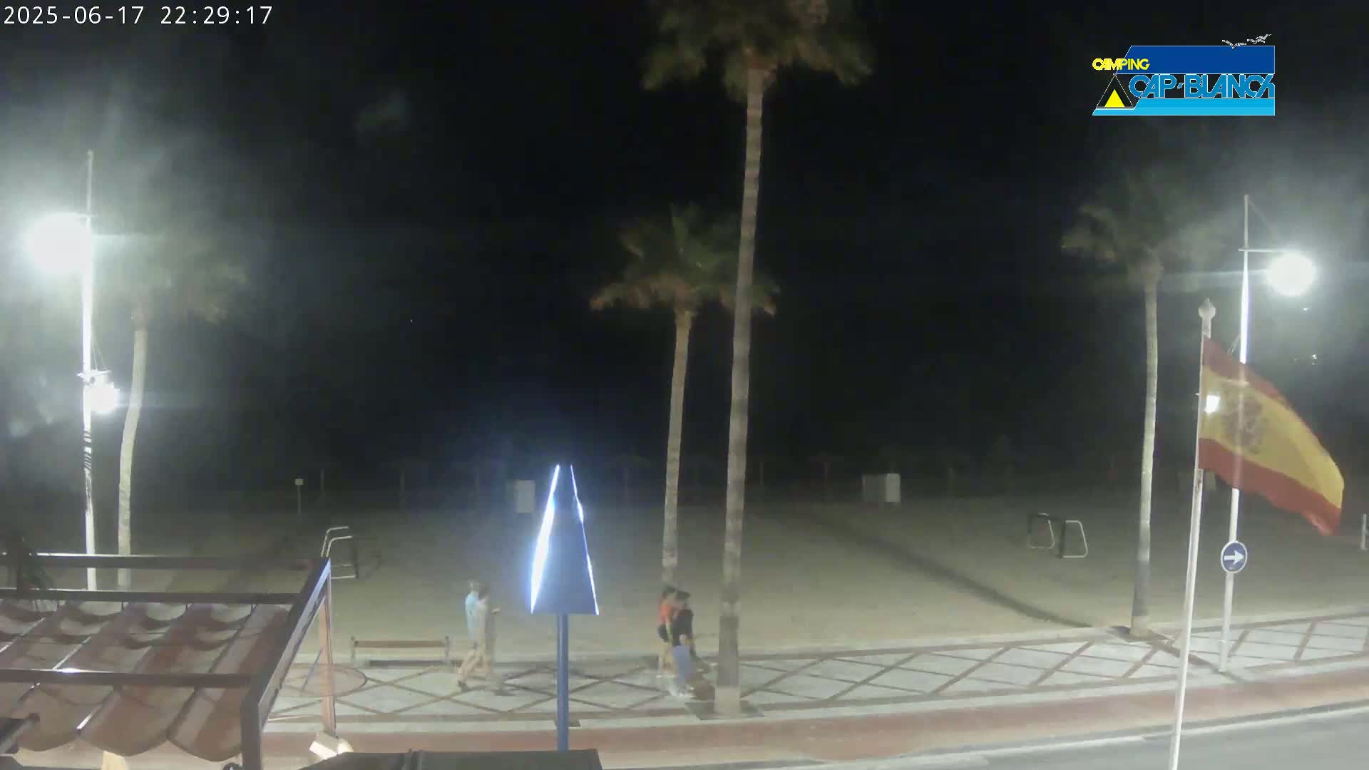 A nighttime view of a sandy beach area with palm trees, a few people walking, and a Spanish flag waving gently in the breeze.