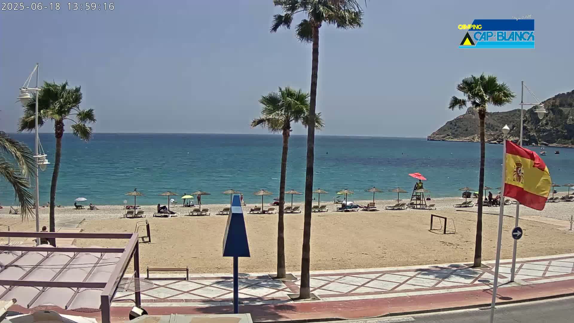 A sandy beach with palm trees, beach umbrellas, and a few people is next to a calm, blue ocean under a clear, sunny sky.