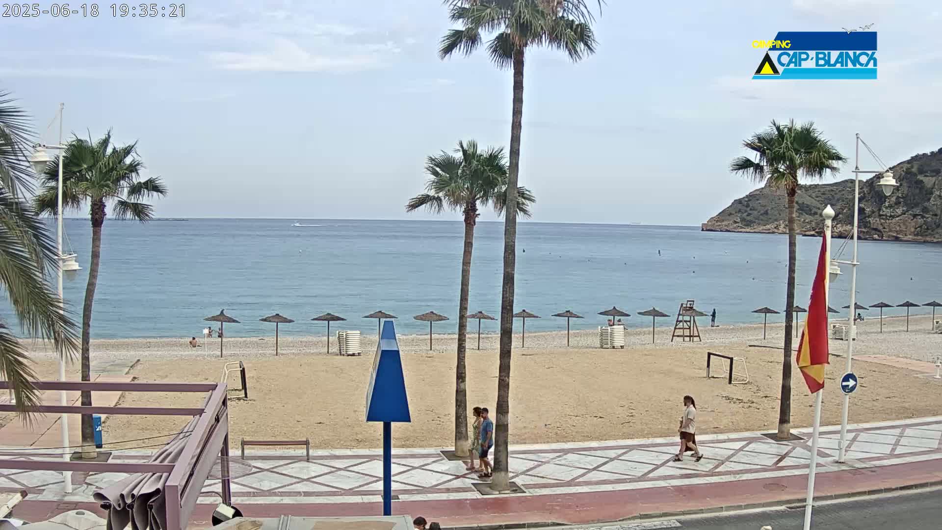 A mostly sunny beach scene shows a sandy beach with several beach umbrellas and a calm blue sea under a mostly clear sky, with palm trees lining the beach and a couple walking along a nearby paved walkway.