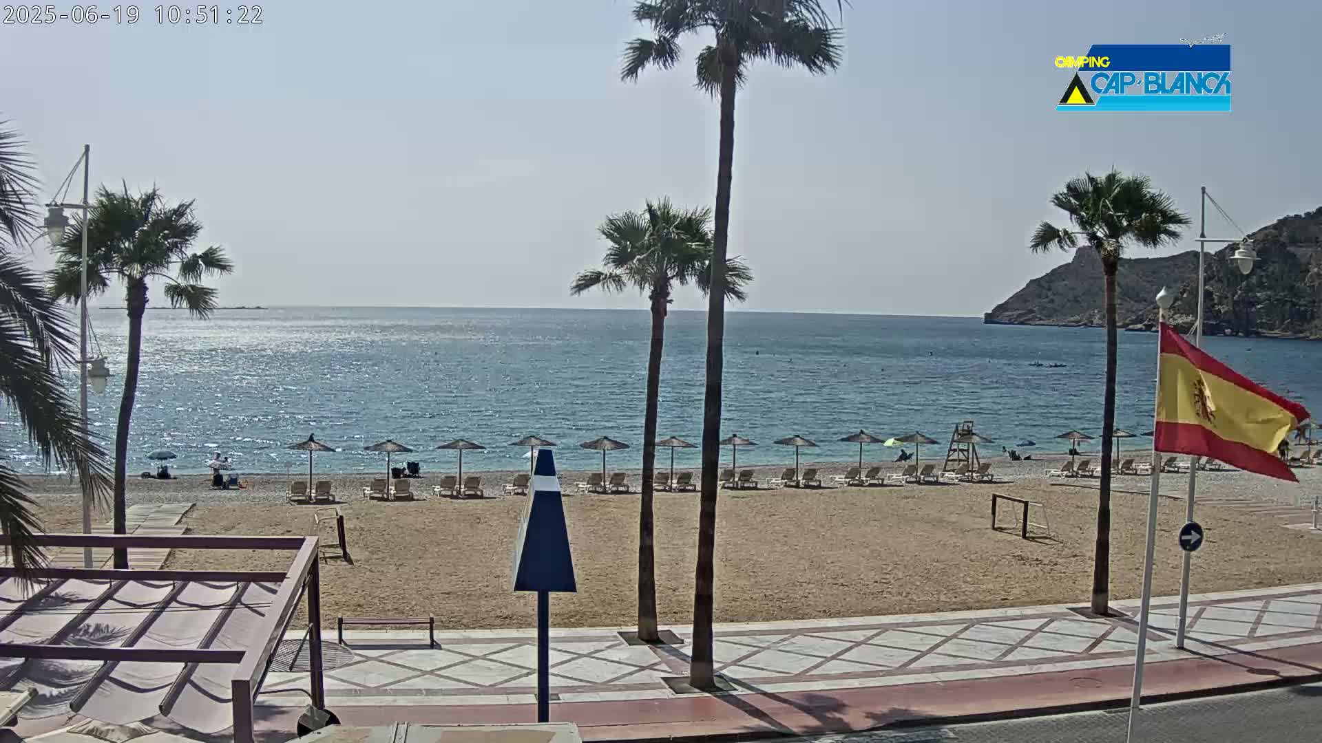 A sunny beach scene shows a sandy shore with beach umbrellas and lounge chairs, palm trees, a calm ocean, and a Spanish flag, all under a clear sky.
