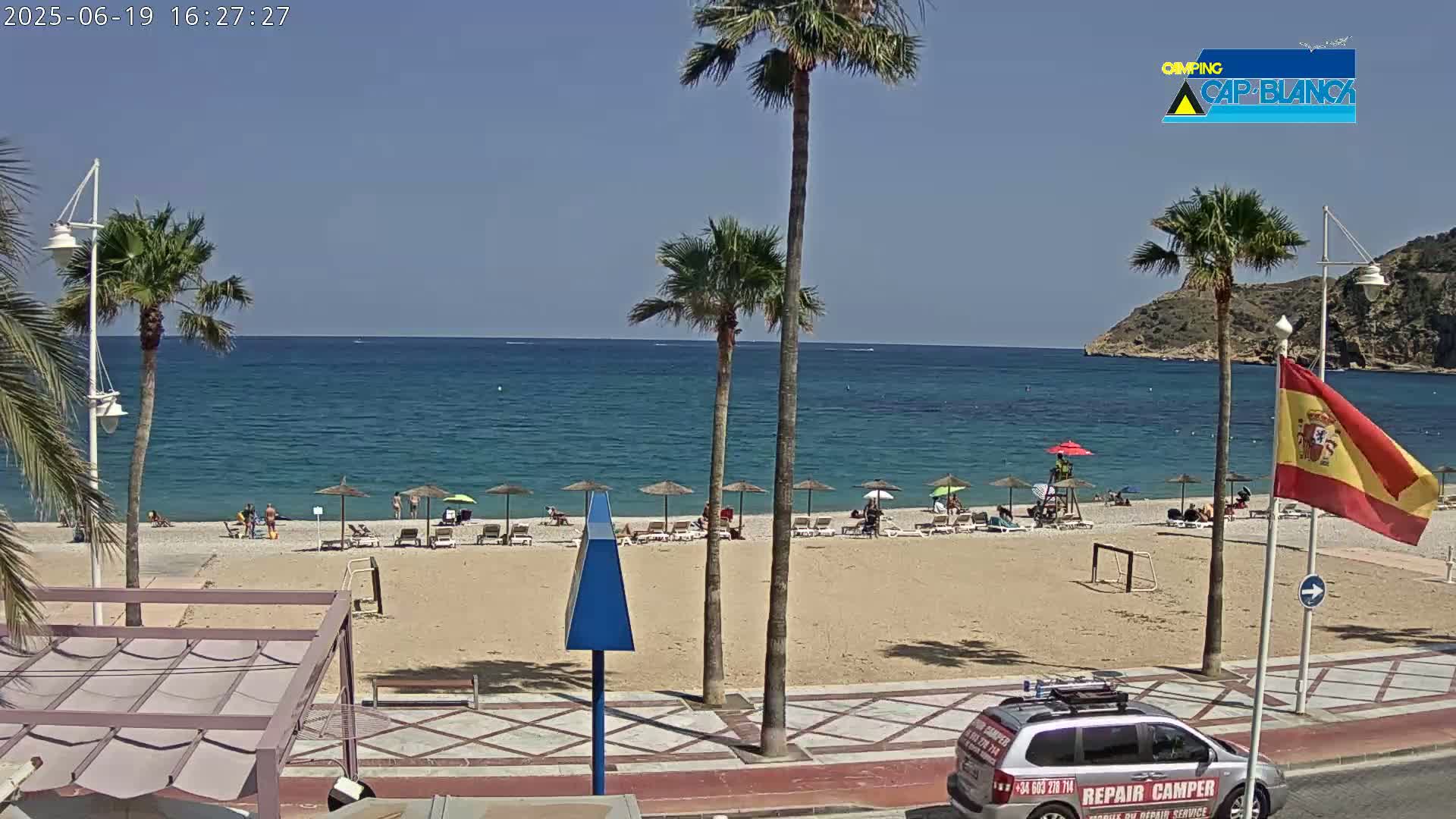 A sandy beach with people relaxing under umbrellas, palm trees, and a calm blue sea under a clear sunny sky.
