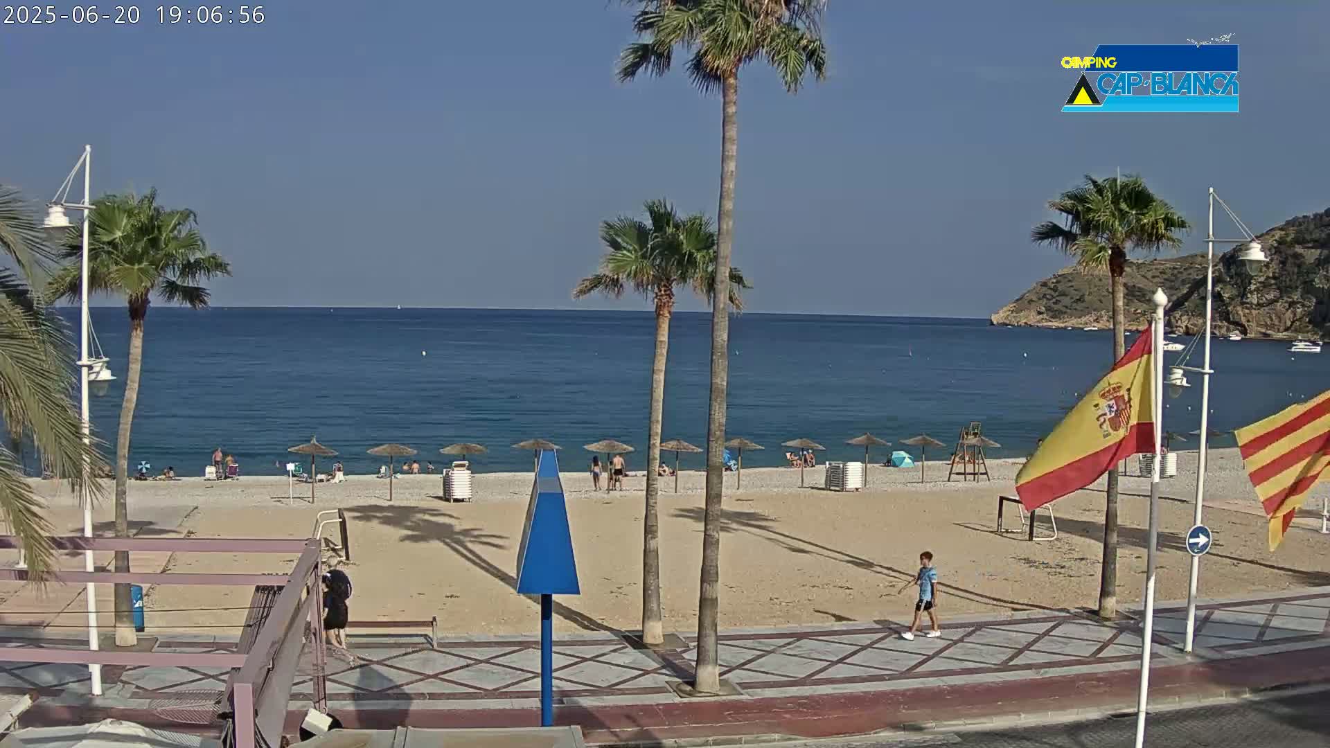 A sandy beach with palm trees and a few people on it, next to a calm blue sea under a clear sunny sky.