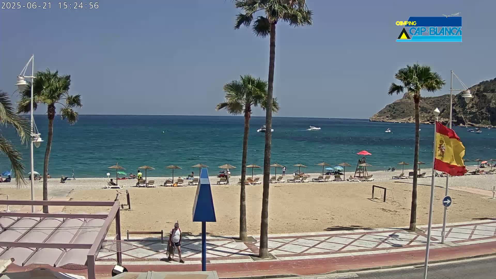 A sunny day at a sandy beach with palm trees, beach umbrellas, and people relaxing, next to a calm blue ocean and a hillside in the background.