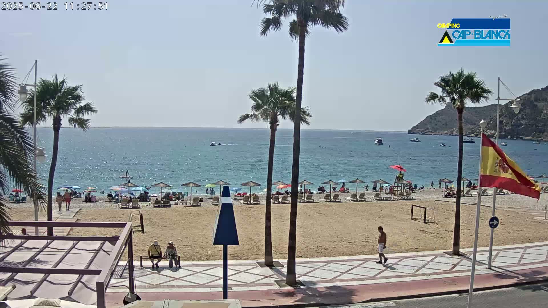 A sandy beach with numerous umbrellas and sunbathers is visible, adjacent to a paved walkway and palm trees, all under sunny skies and overlooking a calm sea.
