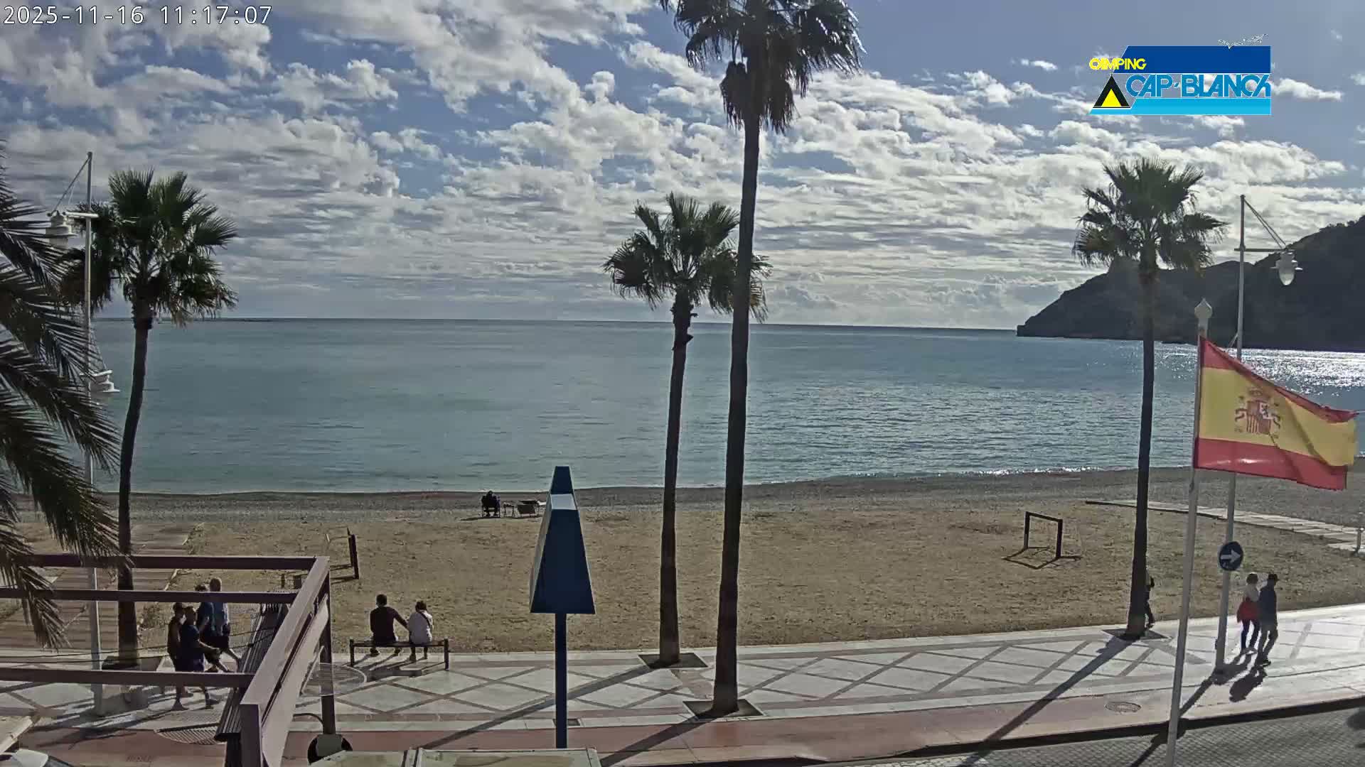 The image displays a sunny, partly cloudy day over a calm turquoise sea and a sandy beach, featuring several palm trees, a promenade with people walking and sitting, and distant hills.