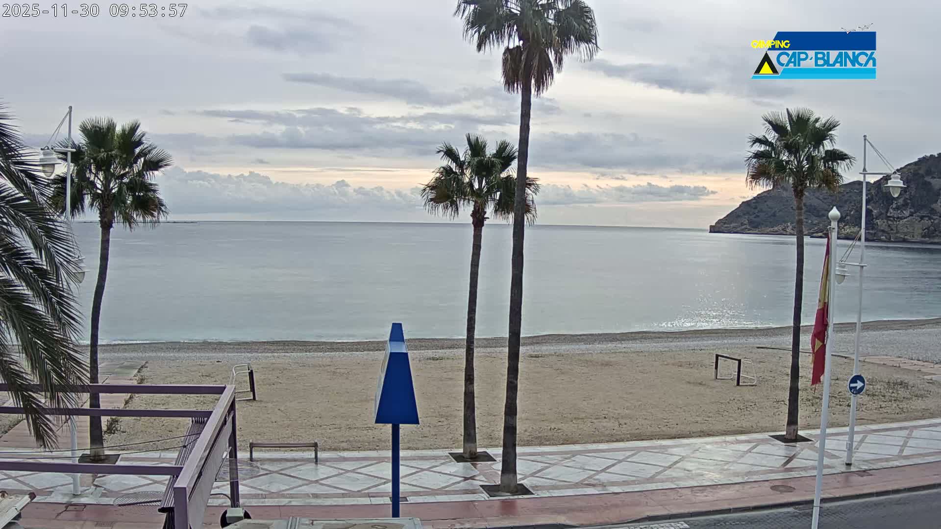 A cloudy day overlooks a calm, pebbled beach lined with palm trees and a promenade, with tranquil sea waters stretching towards distant hills.