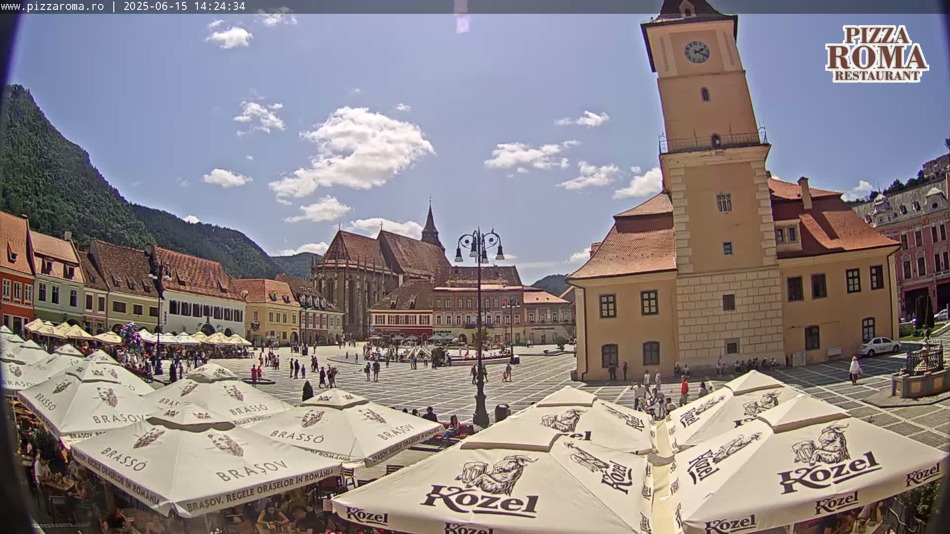 A sunny day in a European town square, filled with people sitting under umbrellas at outdoor cafes, with historic buildings and a leaning clock tower surrounding the cobblestone plaza.