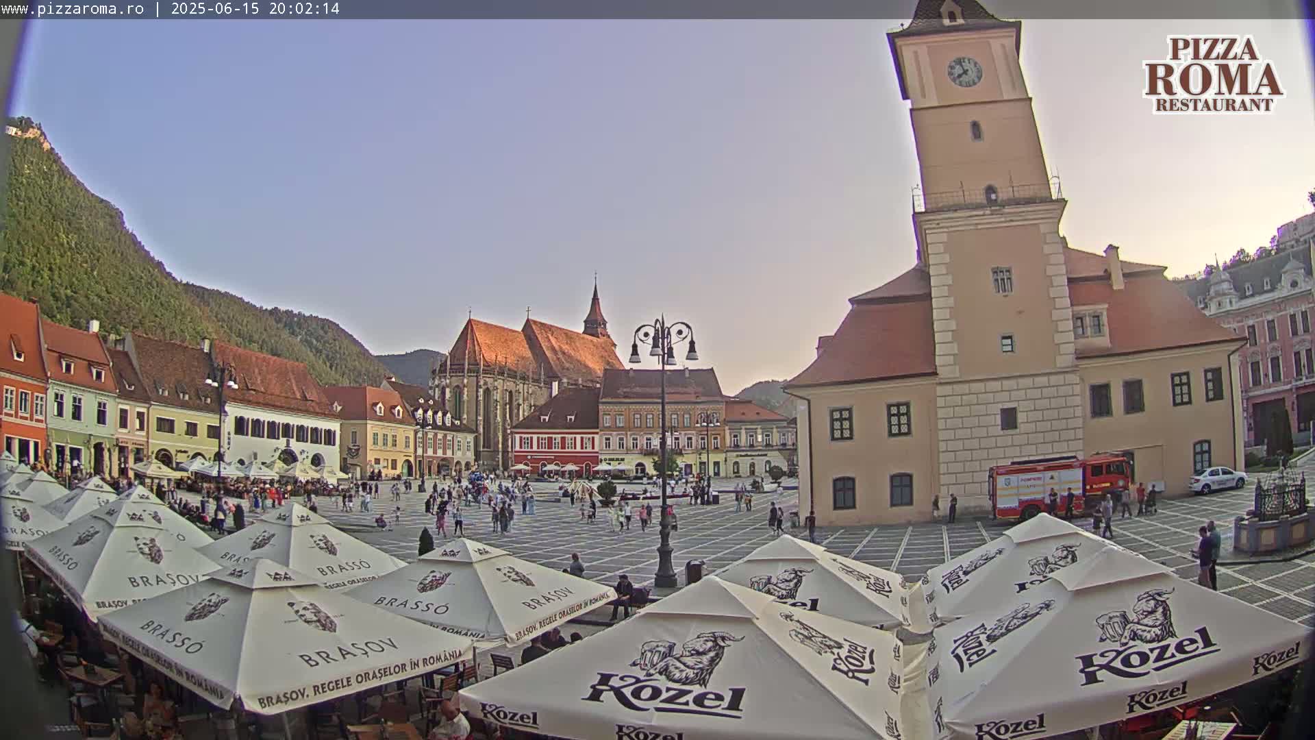 A town square with several buildings, a church, and numerous outdoor cafe umbrellas under a clear, twilight sky.
