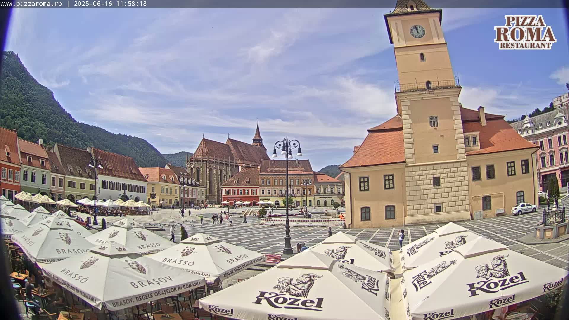 A town square with several outdoor cafes under branded umbrellas, surrounded by historic buildings, including a leaning tower, on a partly cloudy day.