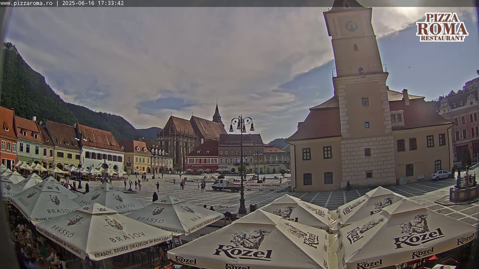 A partly cloudy day reveals a town square with several buildings, including a tall clock tower, surrounded by outdoor seating under branded umbrellas.