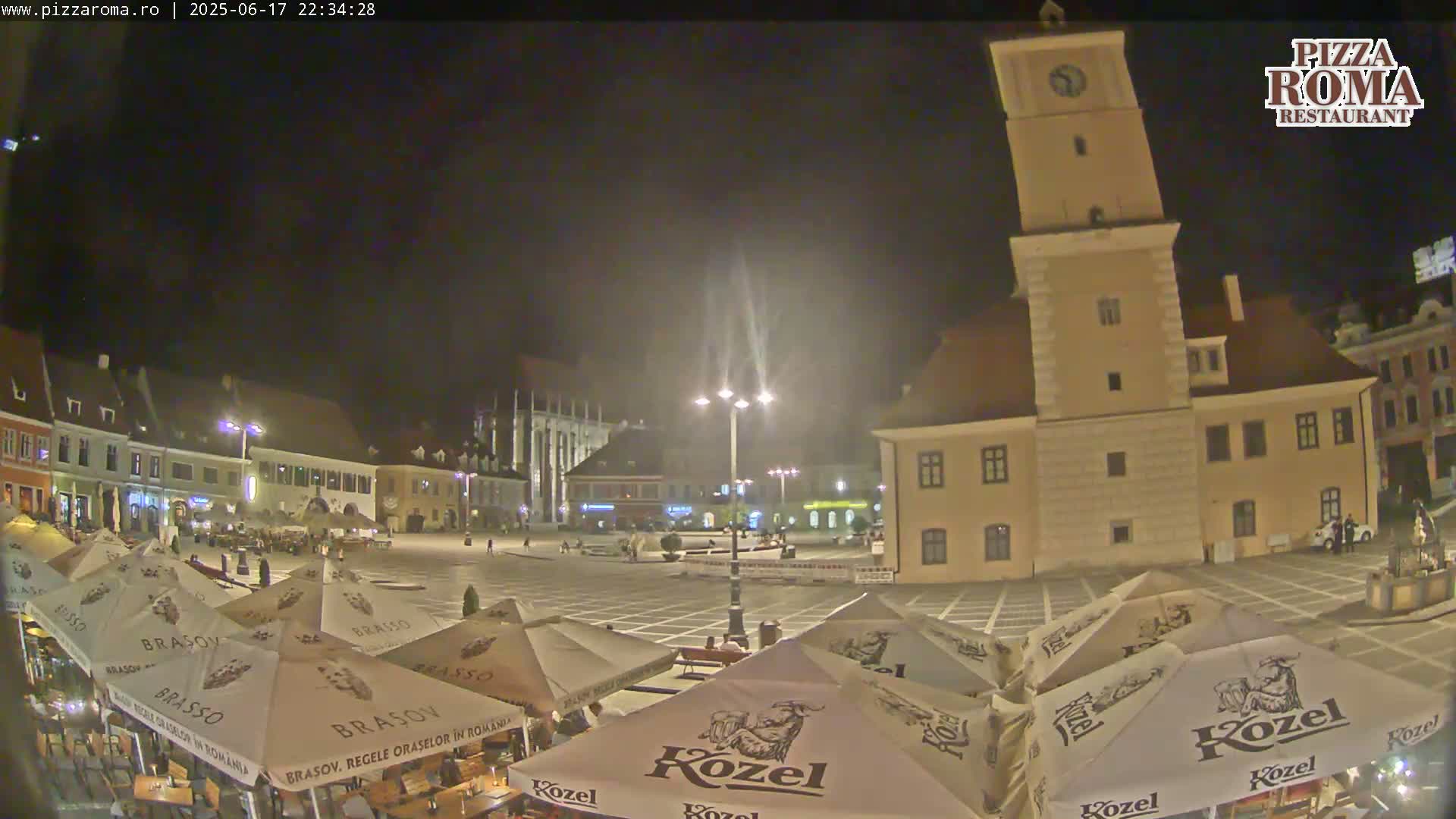 A nighttime view of a town square with several buildings, including a tall clock tower, and outdoor seating under umbrellas.